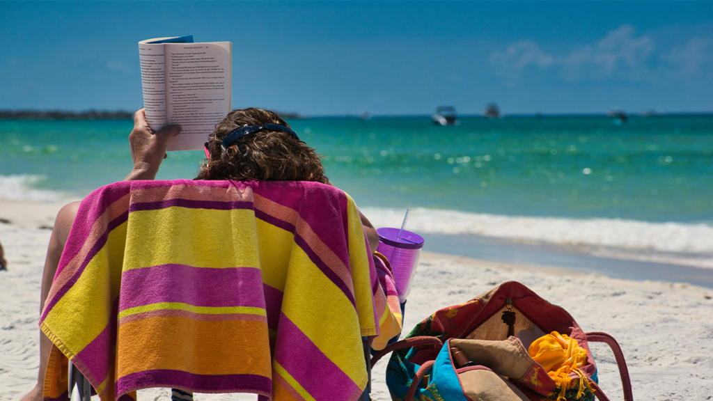 A woman reading a book on a beach chair in front of a teal green ocean with a colorful towel draped on the back.